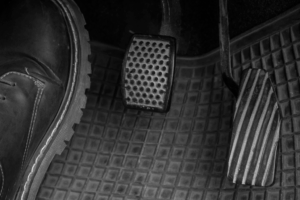 Mecânico a inspecionar o sistema de embraiagem de um automóvel, focando-se no pedal e nos componentes associados a vibrações.