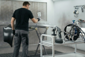 Técnico a utilizar uma pistola de pintura a bateria e airless numa oficina de chapa e pintura, com cabos e equipamentos visíveis.