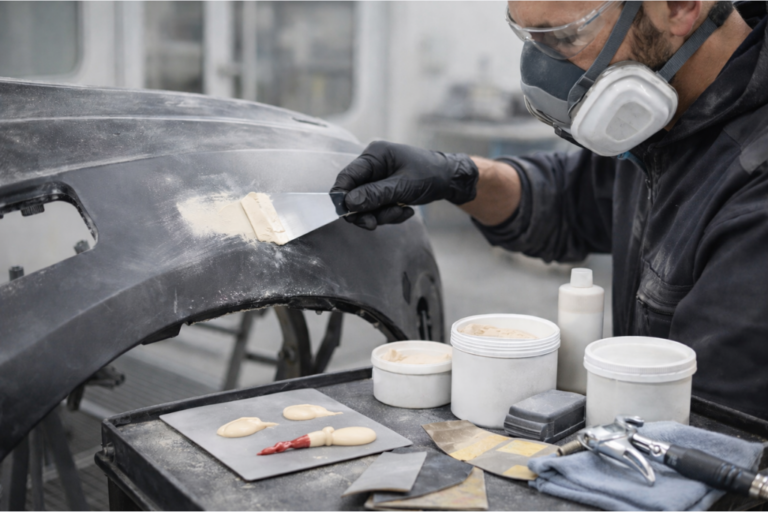 Carrossier appliquant du mastic plastique sur un pare-chocs poncé, avec spatule, abrasifs et produits de préparation sur l’établi, illustrant une réparation durable.