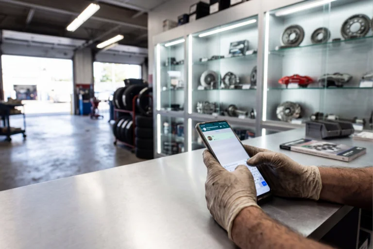 WhatsApp Business para Oficinas Mecânicas: Como Gerir Marcações, Orçamentos e OR num Só Canal 1 Consultor de serviços de oficina automóvel a comunicar com cliente via WhatsApp Business no smartphone com ordem de reparação digital em tablet e baias de serviço visíveis em fundo