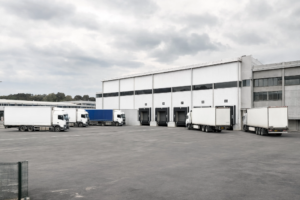 Vue extérieure d'un grand entrepôt logistique blanc avec des quais de chargement numérotés. Plusieurs camions avec remorque blancs et un bleu sont stationnés sur la cour en asphalte, certains étant connectés aux portes de chargement sous un ciel nuageux.
