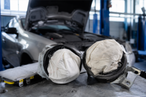 Vue rapprochée de deux airbags frontaux déclenchés ou repliés dans leurs modules de support, posés sur un établi métallique de garage. En arrière-plan flou, on distingue une voiture grise avec le capot ouvert sur un pont élévateur. Parfait pour illustrer des articles sur la sécurité routière, les pièces de rechange de carrosserie, le système SRS ou les réparations après collision.