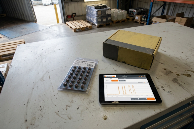 Joints de Soupapes VW 2.0 TDI : Consommation d'Huile, Diagnostic et Prix B2B FR 9 Vue en plongée d'un établi d'atelier métallique sur lequel reposent un blíster de joints de queue de soupape pour moteur VAG 2.0 TDI, une boîte d'emballage en carton jaunie et une tablette numérique affichant des graphiques de diagnostic ou de suivi sur l'application Recambiofacil. L'arrière-plan montre une zone logistique avec des palettes en bois et des fûts.