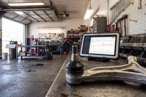 Brazo de suspensión de aluminio con rótula en primer plano sobre un banco metálico con una tablet de diagnosis y el taller de fondo.