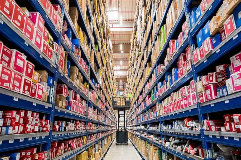 Pasillo central de un almacén industrial con estanterías altas llenas de cajas de piezas de repuesto y un carretillero trabajando