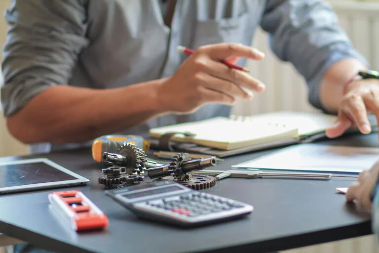 mesa de trabajo oscura y ocupada, enfocada en la discusión. Las manos de una persona en primer plano gesticulan y sostienen un lápiz rojo sobre un cuaderno abierto. El primer plano de la mesa está lleno de herramientas de precisión y piezas de ingeniería: un calibrador vernier