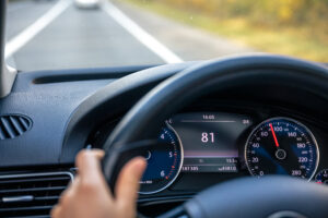Primer plano del interior de un coche en movimiento desde la perspectiva del conductor. Muestra una mano en el volante de cuero. El cuadro de instrumentos digital central muestra la velocidad "81" km/h, la hora "16:05" y una temperatura de "15.5 °C". A la derecha, un velocímetro analógico. A través del parabrisas, se ve una autopista de varios carriles.