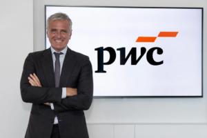 Ejecutivo sonriente en traje posando de brazos cruzados delante del logotipo de la firma PwC en una pantalla.