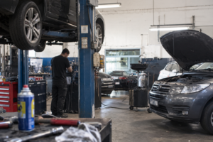 Mécanicien travaillant dans un atelier automobile à côté d'une voiture sur pont élévateur et d'un véhicule capot ouvert.