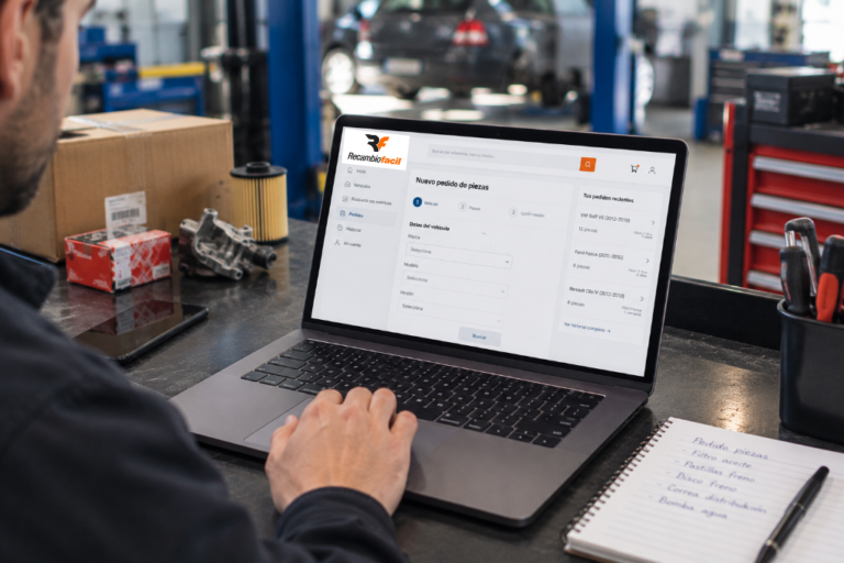 Mecânico a utilizar um computador portátil na oficina para realizar uma nova encomenda de peças no portal Recambiofacil, com peças e um bloco de notas ao lado.